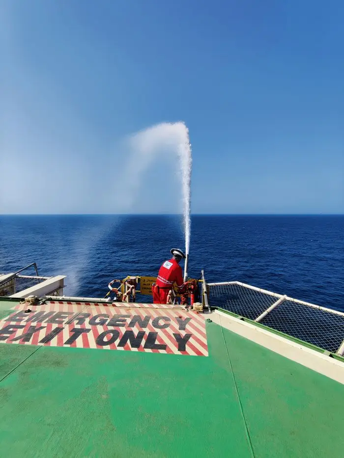 A rescuer conducts a water jet drill on an offshore vessel's deck under a clear blue sky.