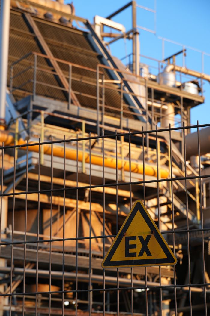 Close-up of a safety sign on a fence in front of an industrial structure at sunset.