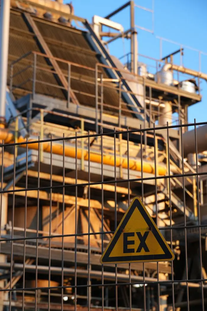 Close-up of a safety sign on a fence in front of an industrial structure at sunset.